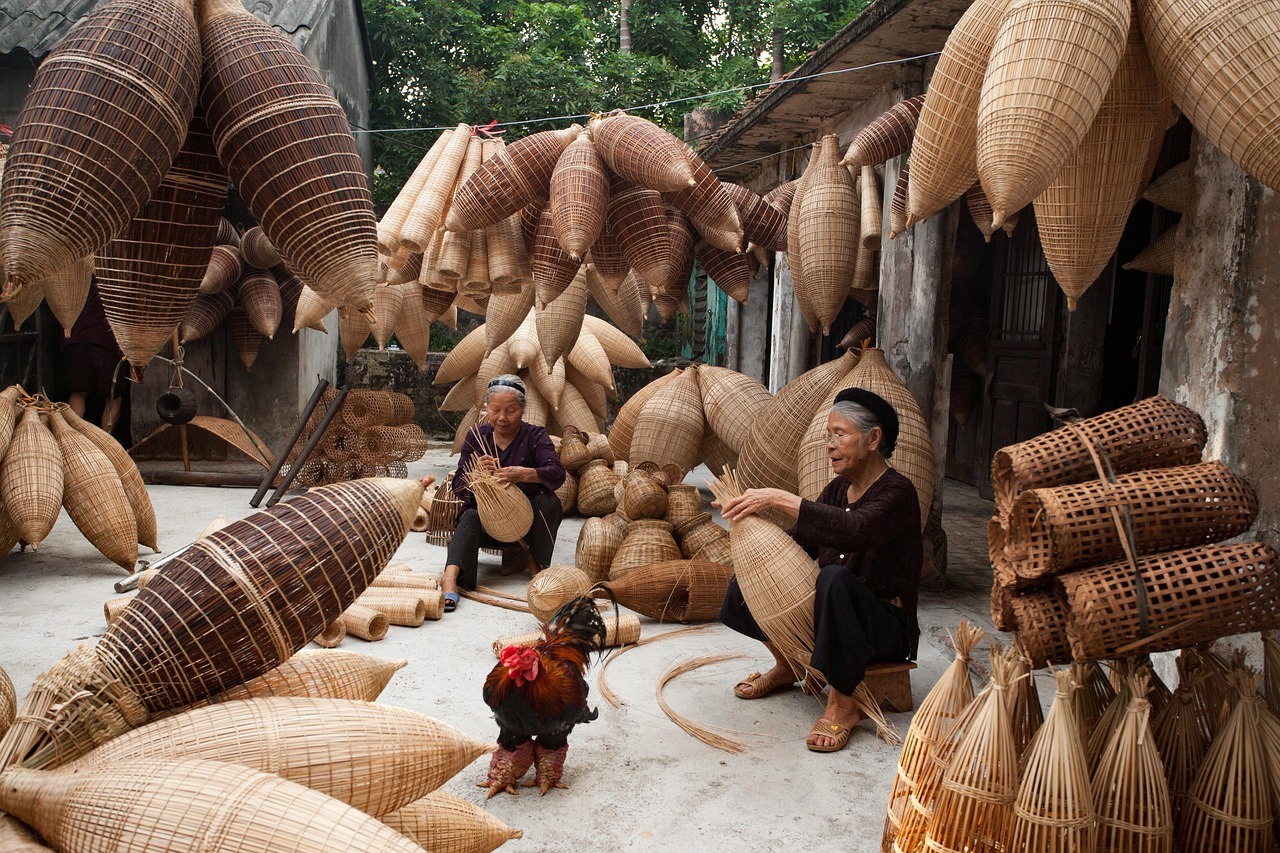 Two seated makers work among many large woven forms in a narrow outdoor workshop.
