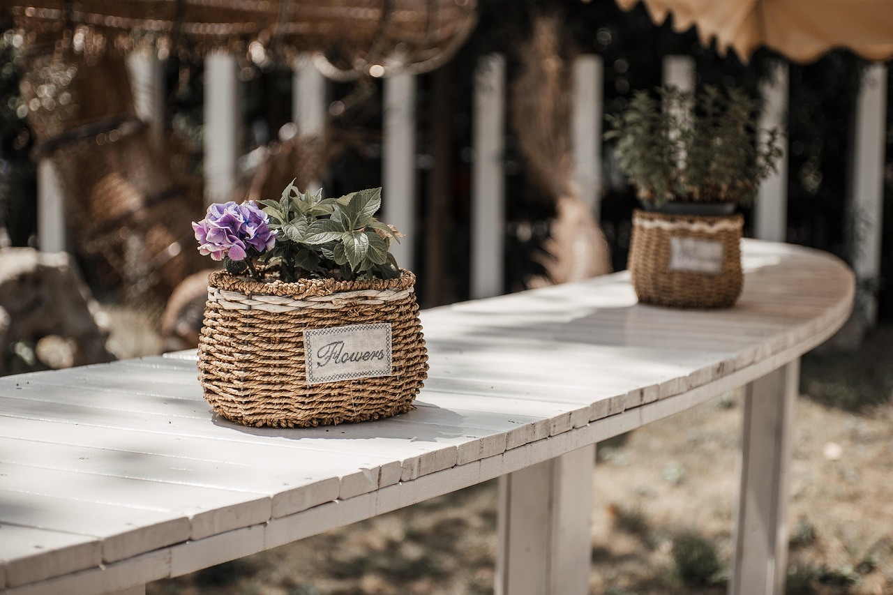 A small flower basket sits on a pale outdoor table with another woven planter blurred behind it.